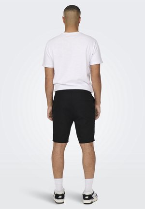 T-shirt bianco in cotone da uomo abbinato a pantaloni shorts neri. Gli shorts hanno una tasca sul retro; indossati con sneakers bianche e nere e calze bianche.