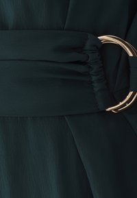 Mørkegrønt stofbælte med en elastisk sektion, der er samlet, fastgjort med en skinnende guldfarvet rund metalspænde på matchende beklædning.