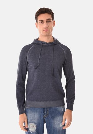 Uomo che indossa una felpa con cappuccio grigio scuro con coulisse e jeans chiari strappati, in piedi contro uno sfondo bianco uniforme.