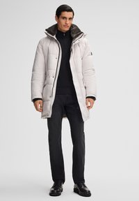 Cappotto imbottito grigio chiaro con collarino foderato in pelliccia, dettagli in zip nera e chiusure a pressione, abbinato a jeans neri e stivali neri lucidi.