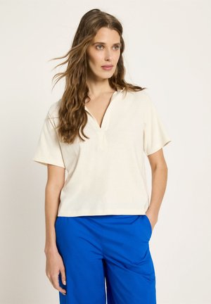 Frau mit langen, welligen braunen Haaren, die ein cremefarbenes Kurzarmshirt und leuchtend blaue Hosen trägt, steht mit einer Hand in der Tasche.