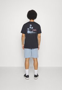 Schwarzes Baumwoll-T-Shirt mit einem blauen Palmenmotiv auf dem Rücken, kombiniert mit hellblauen Shorts und schwarzen Sneakers mit weißen Akzenten.