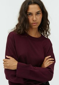 Pull bordeaux à poignets et encolure côtelés, avec une coupe décontractée et un tissu texturé. Le modèle a les bras croisés, mettant en valeur le détail des manches.