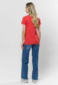 Sarkana īsām piedurknēm t-kreklos ar pieguļošu griezumu, apvienota ar gaiši ziliem platājiem džinsiem un baltām kedām, skatīta no aizmugures.