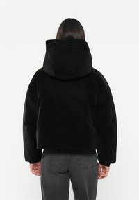 Giacca nera con cappuccio dal design corto, realizzata in un tessuto liscio. Presenta maniche ampie e un colletto alto, indossata su jeans grigi.
