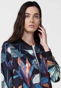 Giacca bomber floreale con sfondo scuro e vivaci foglie tropicali in verde, blu e rosso. Chiusura frontale con zip e polsini a coste.