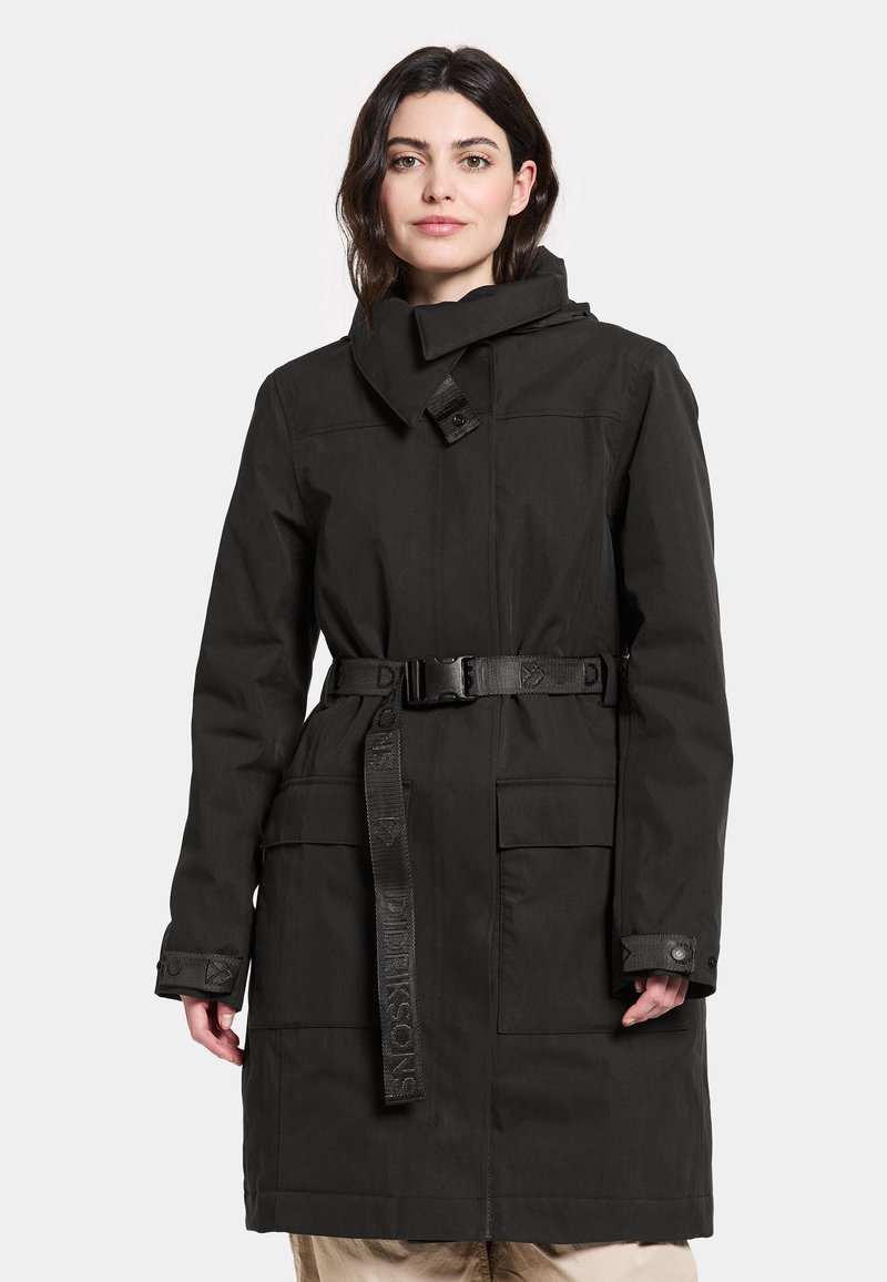Schwarzer langer Mantel aus einem glatten, strapazierfähigen Stoff. Verfügt über einen hohen Kragen, eine taillierte Gürtellinie und zwei Vordertaschen mit Knopfverschluss.