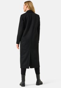 Cappotto lungo nero realizzato in un tessuto liscio, con una fenditura singola sulla schiena, indossato sopra pantaloni scuri e stivali neri alla caviglia.