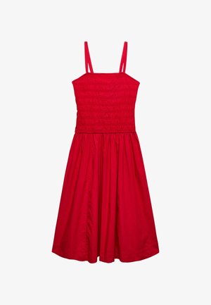 Kvinde iført rød ærmeløs midikjole, cremefarvet cropped jakke, rød vævet clutch og sorte sandaler med røde og hvide flettede stropper.