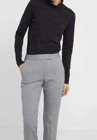 Persoon in een zwarte blouse met lange mouwen, gestoken in een hooggerokte grijze pantalon, staand met één hand in de zak.