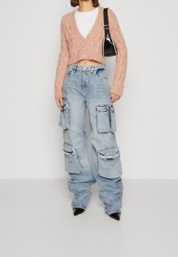 Žena v krátkém růžovém pleteném svetříku, bílém topu, volných světle modrých cargo džínách s velkými kapsami, černých špičatých botách a přes rameno zavěšené tašce.