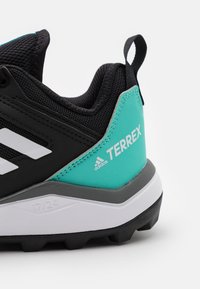 Zapatilla deportiva negra con acentos turquesa, que cuenta con una parte superior de malla texturizada, rayas blancas y una suela gris resistente con lugs de tracción.