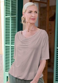 Korte mouwen, gedrapeerde blouse van beige stof, met een ontspannen pasvorm en een zachte textuur. Geplaatst tegen groene luiken voor contrast.