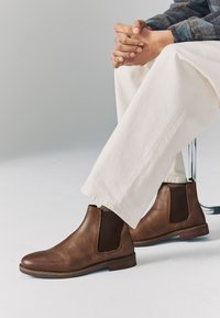 Bottes Chelsea en cuir marron avec panneaux latéraux élastiques, coutures détaillées et petit talon, associées à un pantalon large crème.