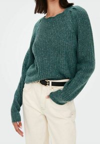 Pull en maille côtelée vert avec des manches longues, un col rond et une coupe décontractée, associé à un pantalon couleur crème et une ceinture noire.
