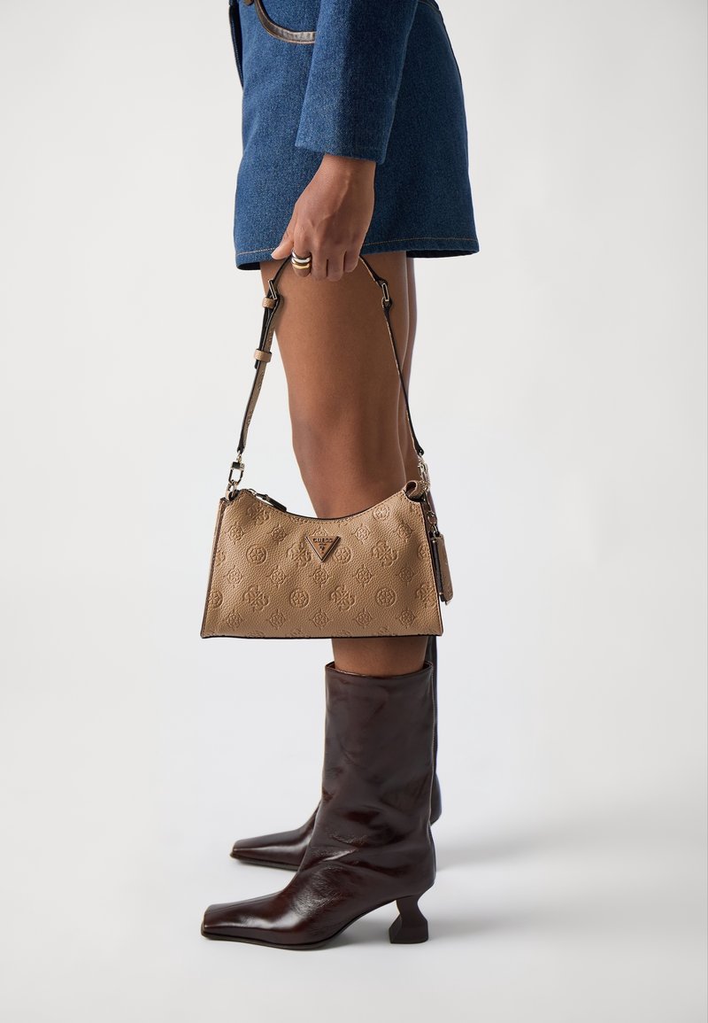 Femme portant une jupe bleue et des bottes marron foncé mi-mollet, tenant un sac à main beige texturé avec un motif et une bandoulière réglable.