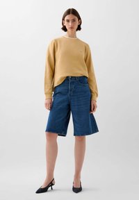 Geel crewneck sweatshirt, blauwe denim bermuda shorts en zwarte hakken. De stof lijkt glad; de shorts hebben een losse pasvorm met een opgerolde zoom.