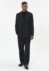 Čierny dvojradový blejzer a voľné nohavice, vyrobené z hladkej látky. Nosí sa s čiernym rolákom a lesklými čiernymi topánkami.