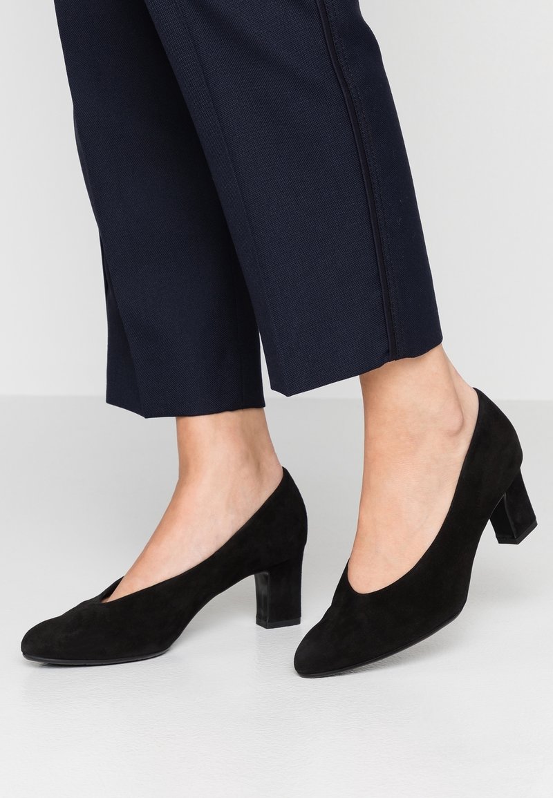 Pieds portant des chaussures à talon bloc en daim noir associées à un pantalon court bleu marine foncé sur un fond clair.