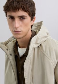 Young man with short brown hair wearing a beige hooded jacket and brown shirt, looking directly with a neutral expression on gray background.