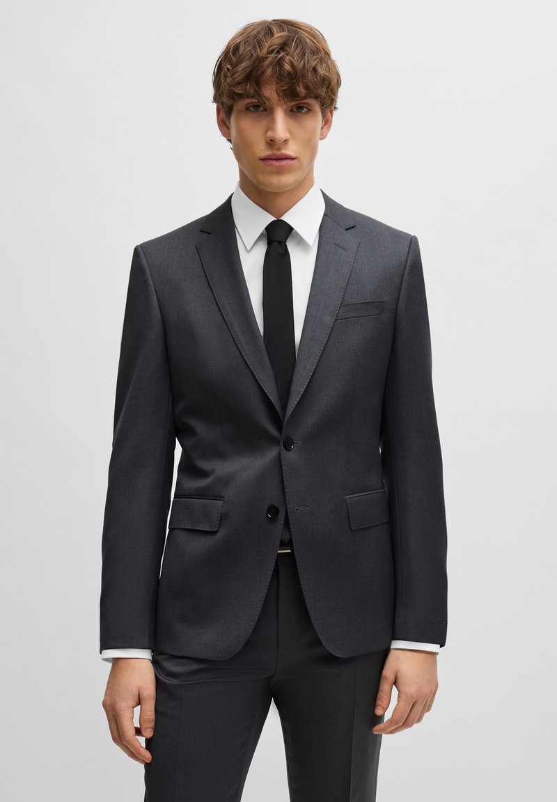 Donkergrijs pak met een gestructureerd ontwerp, enkelgeknoopte blazer, notched revers en een twee-knoopsluiting. Vergezeld van een wit overhemd en een zwarte stropdas.