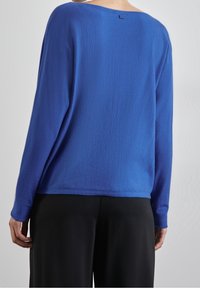 Blue long-sleeve sweater with a relaxed fit, textured fabric, wide neckline, and small logo tag at the back, paired with black pants.