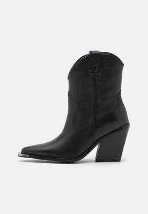 Sort læder cowboystøvle med spids tå, metal tåhætte, dekorative syninger, trækflap og skråt blokhæle mod en ensfarvet baggrund.