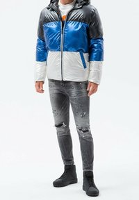 Gepolsterte Jacke mit schwarzen, blauen und weißen Paneelen, Reißverschluss und Kapuze; getragen mit ausgewaschenen grauen Jeans und schwarzen High-Top-Schuhen.