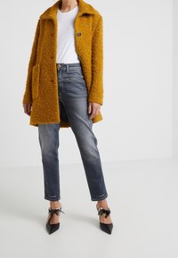 Abrigo texturizado mostaza con cierre de botón, combinado con jeans rectos de tiro oscuro y tacones negros de punta con lazos en el tobillo.