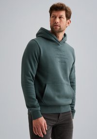 Grüne Kapuzenjacke aus weichem Material, mit einer Vorderstasche, gerippten Bündchen und geprägtem Text. Kombiniert mit dunklen Jeans.