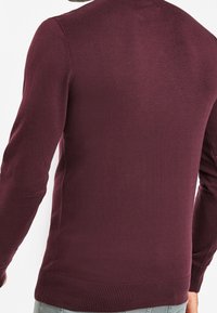 Mann mit einem enganliegenden, dunkelburgunderfarbenen Langarmpullover mit gerippten Bündchen und Saum, von hinten gezeigt.