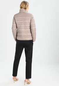Beige pufferjack met een hoge kraag, doorgestikt ontwerp en geribbelde details, in combinatie met zwarte broek en lichtgekleurde schoenen.