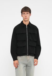 Zwarte corduroyjas met een rits aan de voorkant, voorzien van vier grote voorkruiden, een kraag en mouwen met ribgebreide manchetten. Gekleed met lichte jeans.