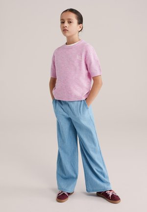 Maglioncino rosa chiaro a maniche corte con finitura strutturata e dettaglio bianco al colletto, abbinato a pantaloni larghi in denim blu e scarpe color marrone scuro.