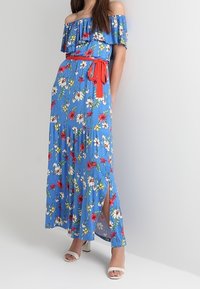 Frau in einem blauen, schulterfreien Maxi-Kleid mit Blumendruck, rotem Gürtel und weißen offenen Schuhen, steht vor einem schlichten, hellen Hintergrund.