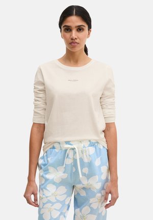 Frau mit dunklem Pferdeschwanz, die ein cremefarbenes Langarmshirt und hellblaue Hosen mit großem weißem Blumenmuster und einem Kordelzug trägt.