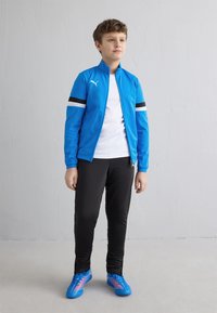 Dječak stoji uz bijeli zid, nosi plavu Puma jaknu, bijelu majicu, crne hlače i plave sportske cipele.