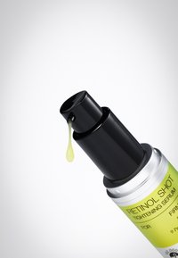 Pompe noire distribuant une goutte de sérum jaune du flacon de sérum raffermissant Retinol Shot avec une étiquette vert citron sur fond blanc.