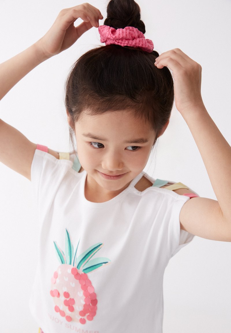 Camisa de manga corta blanca con un diseño de piña rosa. Accentos en los hombros en colores pastel. Niño/a lleva una diadema arrugada rosa en el cabello.