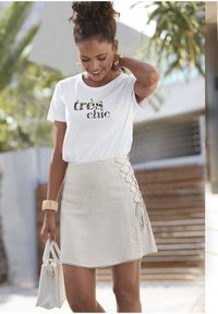 Weißes T-Shirt mit "très chic" in einem strukturierten goldenen Muster bedruckt, kombiniert mit einem beigen Rock mit seitlichen Schnürdetails.