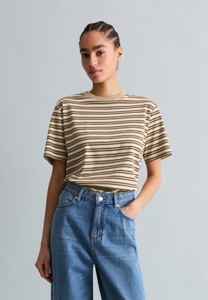 Młoda osoba z plecionymi włosami, ubrana w beżowy T-shirt w paski oraz niebieskie, wysokodrise spodnie dżinsowe, stojąca z jedną ręką w kieszeni.