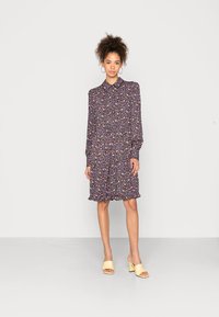 Vestido com estampado floral em púrpura escuro, com mangas longas, colarinho e bainha com babados. Usado com saltos amarelos de bico aberto para contraste.