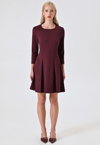 Kleid in A-Linie in Kastanienbraun, aus einem strukturierten Stoff gefertigt, mit einem runden Ausschnitt, Dreiviertelärmeln und gerüschtem Detail am Saum.