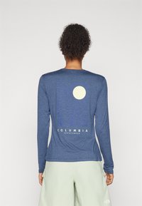 Osoba s kudrnatými vlasy, oblečená v modré košili s dlouhým rukávem Columbia, na které je na zadní straně design měsíce a vlny, a světle zelených šortkách.