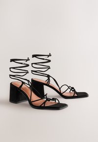 Sandali con tacco in suede nero con punta quadrata, sottili cinturini incrociati e lacci a spirale alla caviglia. Il tacco è un design a blocco robusto.