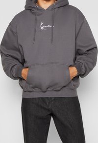 Grå hoodie med en framficka, dragsko på huvan och vit logodetalj. Tyget verkar mjukt, med ribbade mudd och nederkant.