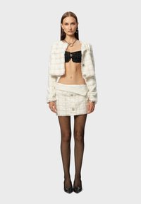 Witte gestructureerde cropped jack over een zwarte gebreide crop top, gecombineerd met een witte minirok en zwarte visnetkousen. Zwarte puntige hakken.