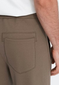 Khaki geklede broek met een back pocket, zichtbaar stikwerk en een aansluitende tailleband. Hand op de heup geplaatst.