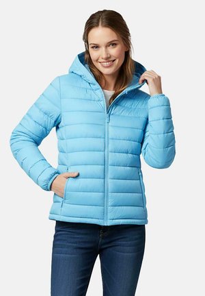 Jonge vrouw die lacht, gekleed in een lichtblauw doorgestikt jasje met capuchon, één hand in de zak, de andere hand die de capuchon aanpast, en een spijkerbroek aan.