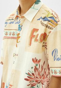 Camisa de manga corta con cuello, hecha de tela ligera, que presenta una base crema con patrones florales y de texto en colores rojo, azul y verde.
